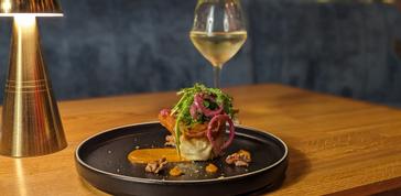 HYDE Hotel | Galway | Stylishly plated gourmet dish topped with greens and pickled onions, served with sauce and nuts on a black plate, paired with a glass of white wine 