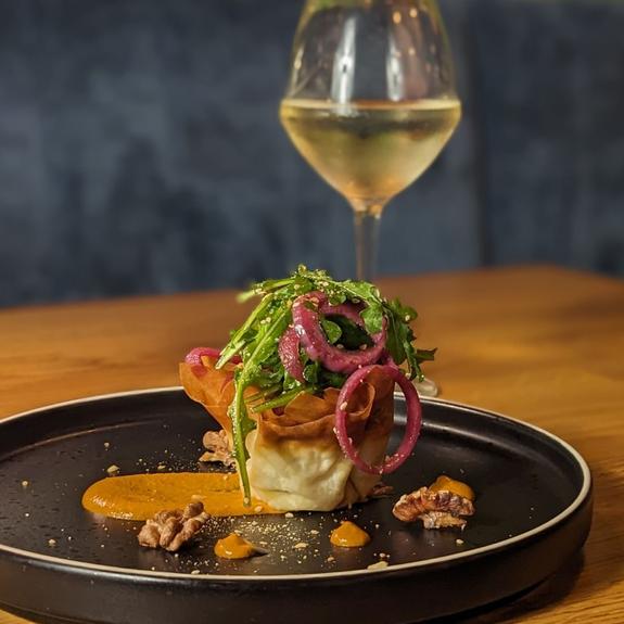 Stylishly plated gourmet dish topped with greens and pickled onions, served with sauce and nuts on a black plate, paired with a glass of white wine 