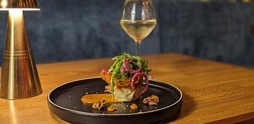 HYDE Hotel | Galway | Stylishly plated gourmet dish topped with greens and pickled onions, served with sauce and nuts on a black plate, paired with a glass of white wine